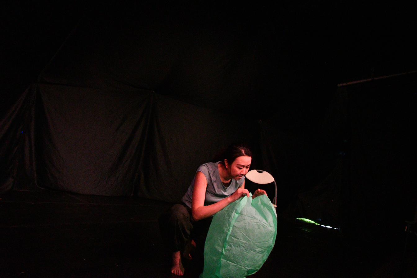 A performer holds an everyday object, hand raised, in Between Strands.