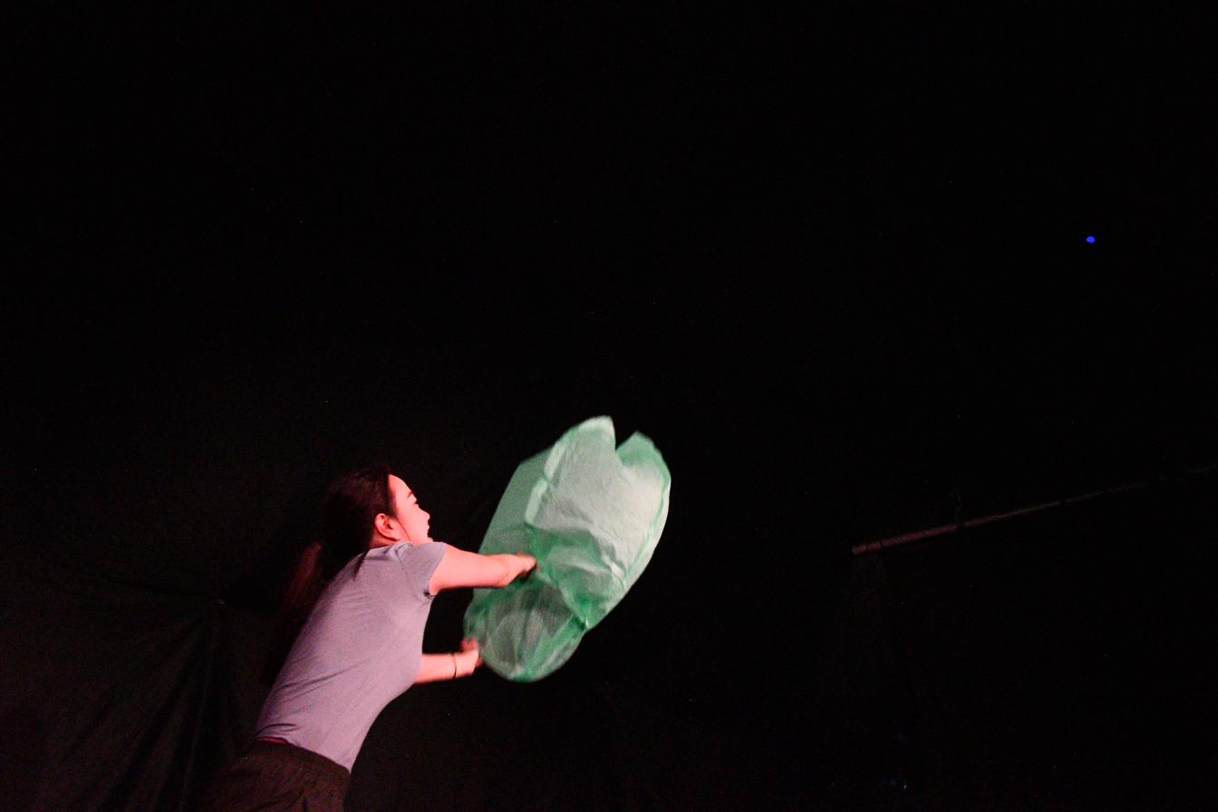 A performer releases a glowing paper lantern into the dark in Between Strands.