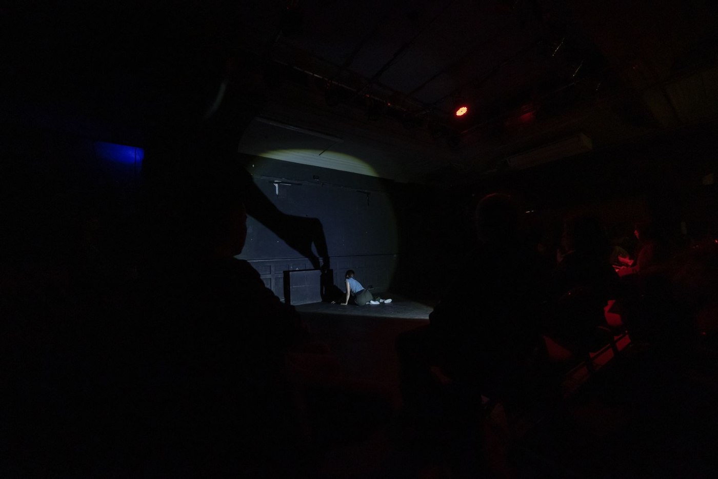 A wide view of the dark studio: a lone performer in a pool of light, audience in silhouette.