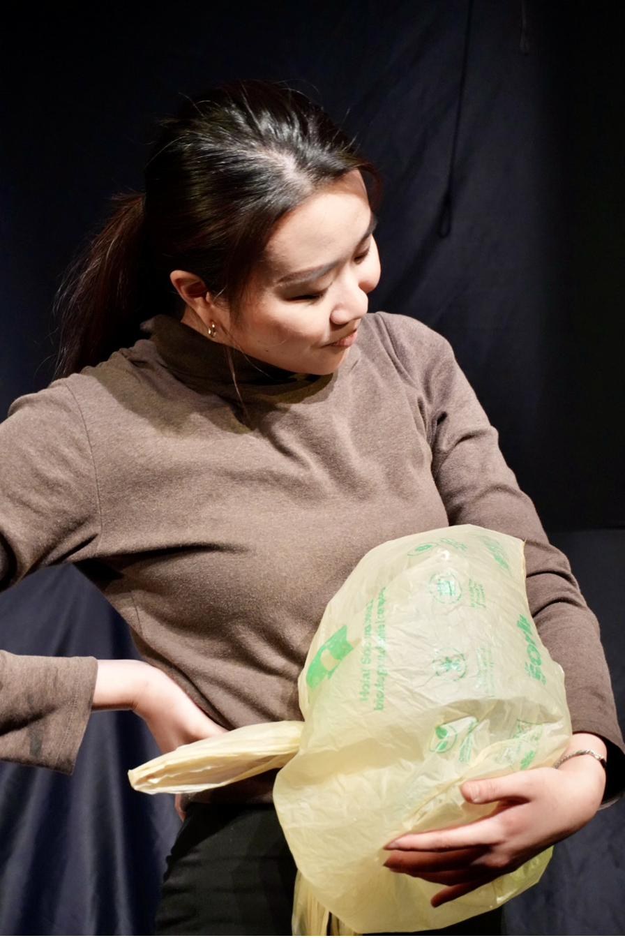 A performer cradles a plastic bag like something precious, lit against black.