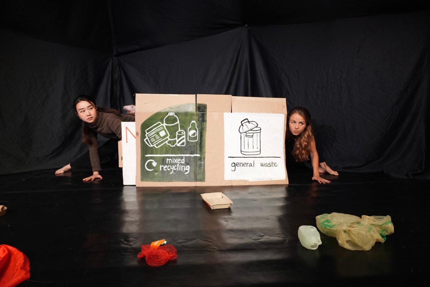 Both performers peer out from behind painted mixed-recycling and general-waste boxes.