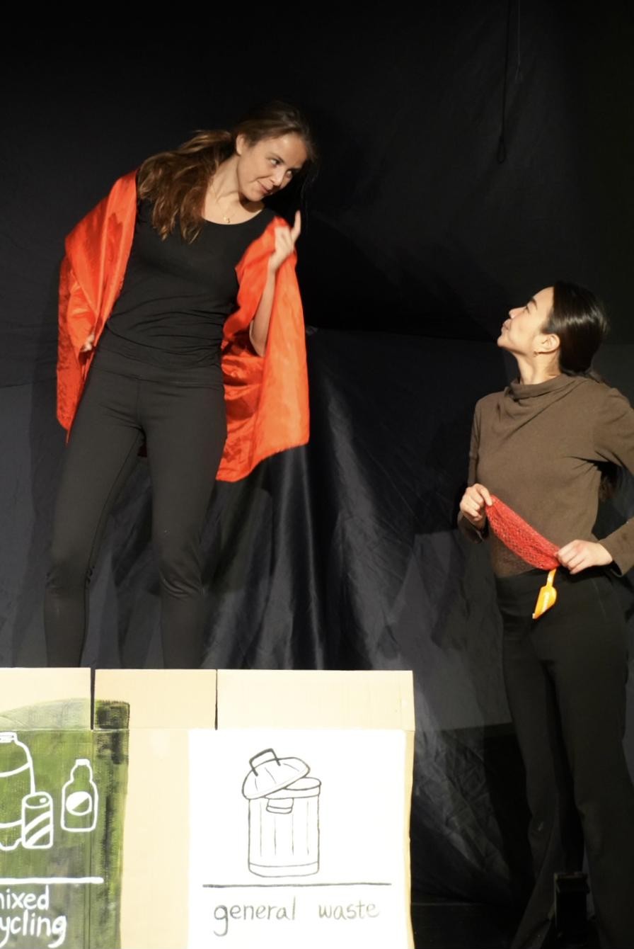 Two performers beside mixed-recycling and general-waste bins on stage.