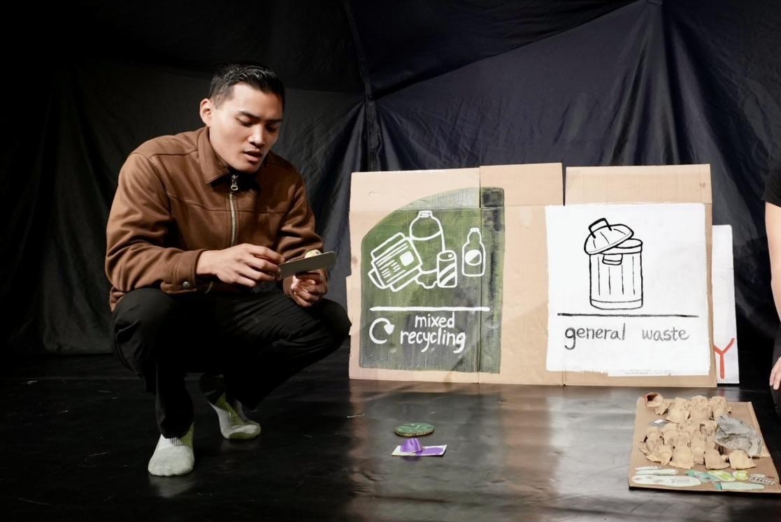A performer crouches to examine a small handcrafted object beside the painted bins.