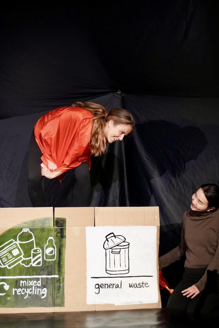 A performer in red leans over the mixed-recycling and general-waste boxes.
