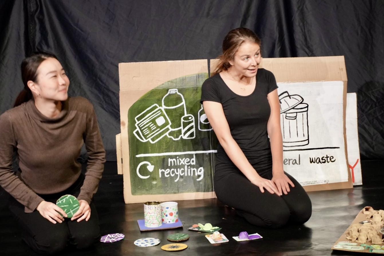 Both performers kneel beside the painted bins, surrounded by small art objects made from waste.